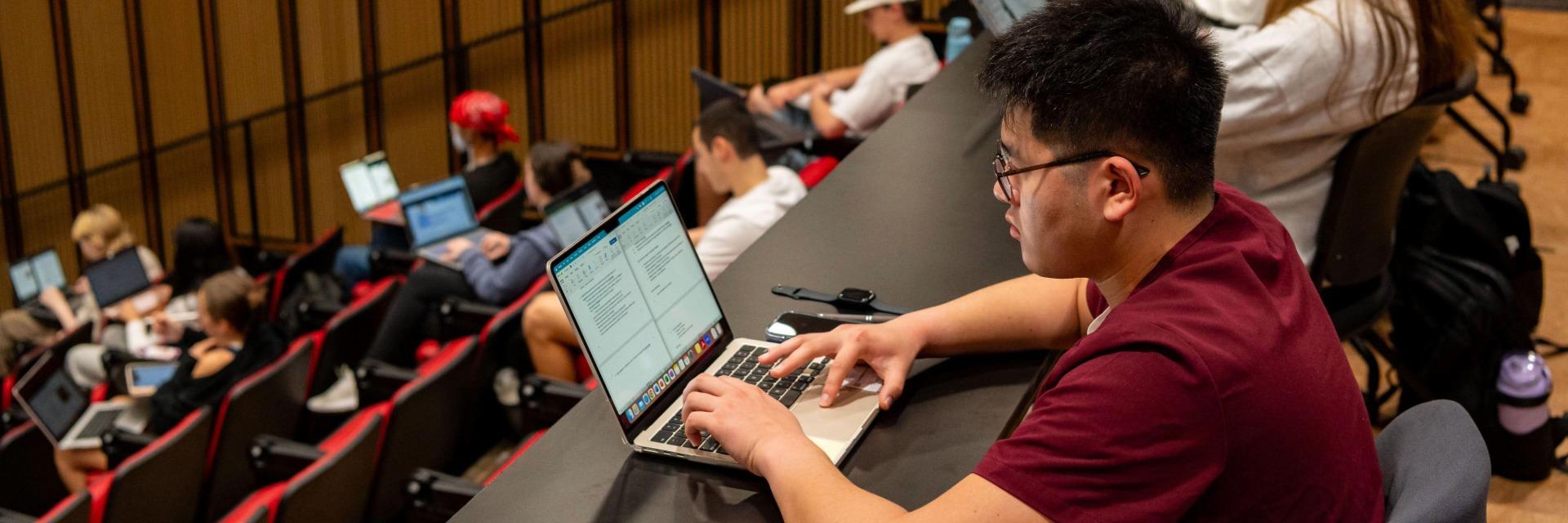 Student in the back row of a lecture hall taking notes on his laptop.